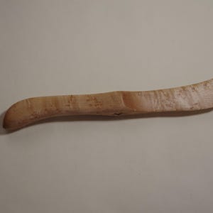 May include: A wooden butter knife with a natural wood grain finish. The knife has a curved handle and a slightly rounded blade.