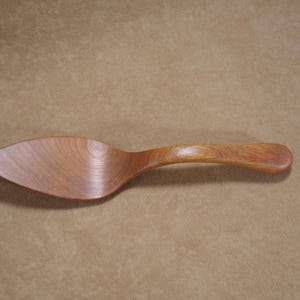 Può includere: Una paletta per torte in legno con un manico curvo e una lama larga e piatta. La paletta è realizzata in legno marrone chiaro e ha una finitura liscia e lucida. La paletta per torte è progettata per servire pasticcini e dessert.
