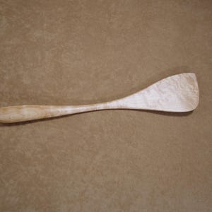 May include: A wooden spatula with a long handle and a wide, curved blade. The spatula is made of light-colored wood, with a natural grain pattern visible throughout. The spatula is designed for cooking and food preparation.