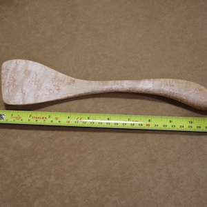 May include: A wooden spatula with a curved handle and a wide, flat head. The spatula is approximately 12 inches long. The wood has a light color with a unique grain pattern. A yellow tape measure is placed next to the spatula.