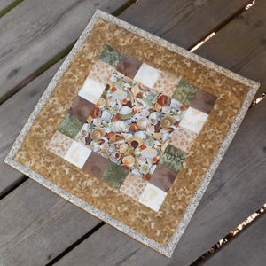 May include: A square quilted placemat with a seashell pattern at the center. The placemat features a patchwork design with squares of brown, green, and white fabric. The outer border is a light brown fabric with a floral pattern, and a white lace trim.