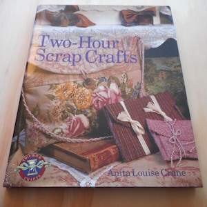 May include: A book titled "Two-Hour Scrap Crafts" by Anita Louise Crane. The cover features images of handmade crafts, including fabric pouches with bows and a floral-patterned bag. The book is on a light-colored surface.