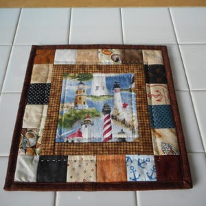 May include: Square quilted coaster with a lighthouse scene in the center. The coaster has a patchwork design with various fabric patterns and colors, including brown, beige, and blue. The border is a solid dark brown.