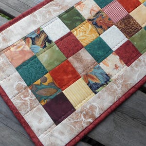 May include: A patchwork table runner with a cream background and a border of red fabric. The runner is made up of squares of different fabrics in shades of brown, green, red, and yellow. The fabrics feature various patterns, including leaves, flowers, and geometric designs.