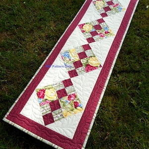 May include: A white table runner with a burgundy border and a floral pattern. The runner is quilted with a leaf design and has a scalloped edge.