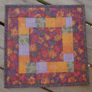 May include: Square quilted placemat with a floral pattern in shades of red, orange, and purple. The center features a geometric design with squares of orange, lavender, and green. The border is a solid purple.