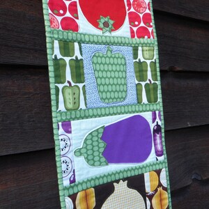 May include: A colorful fabric wall hanging with a patchwork design featuring various fruits and vegetables. The design includes a red tomato, green peppers, a purple eggplant, and a yellow onion.