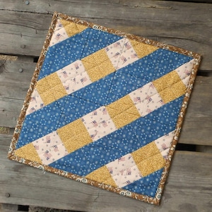 May include: A square quilted fabric mat featuring a geometric pattern of alternating diagonal stripes. The design includes blue fabric with white stars, gold textured fabric, and cream fabric printed with small American flag motifs.