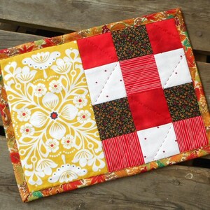 May include: A rectangular quilted fabric mat featuring a yellow panel with a white floral pattern on the left and a patchwork section on the right. The patchwork consists of red, white, and dark floral squares, all bordered by a colourful autumn leaf print.