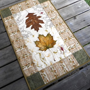 May include: A rectangular table runner with a white background and a brown, green, and gold patterned border. The center of the runner features two large, detailed autumn leaves in shades of brown, yellow, and orange. The leaves are surrounded by a delicate, swirling vine pattern.