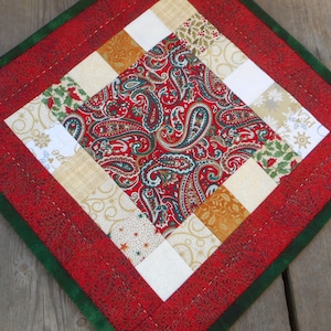 May include: A square, quilted Christmas placemat with a red paisley centre. The placemat features a red border with a green edge. The centre is surrounded by alternating squares of white, gold, and patterned fabric.