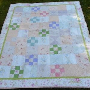 May include: A patchwork quilt with a floral print in shades of pink, green, and white. The quilt is made up of squares and rectangles, and the edges are trimmed with a green border.
