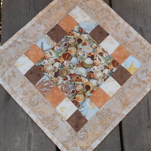 May include: A square quilted placemat featuring a seashell design. The center showcases a variety of seashells, starfish, and coral. The border is a patchwork of brown, orange, and cream squares, with a patterned edge. The overall color palette is warm and beachy.