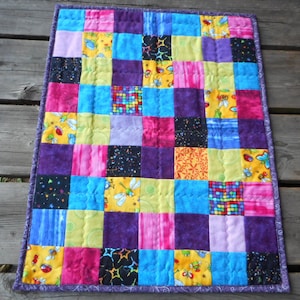 Peut inclure: Une courtepointe colorée en patchwork avec une variété de tissus, dont du violet, du rose, du jaune, du bleu et du noir. La courtepointe est composée de carrés et est matelassée avec un motif simple.