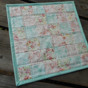 May include: A light blue and white patchwork quilt with a floral pattern. The quilt is made of squares of fabric sewn together.