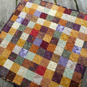 May include: A square patchwork quilt featuring a grid of small fabric squares in shades of yellow, orange, red, purple, green, and cream. The quilt is finished with a dark purple border and displays intricate, swirling machine-stitched patterns.