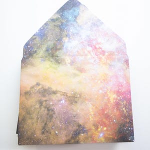 May include: A stack of envelopes with a galaxy print. The print features a swirling nebula with shades of pink, blue, yellow, and orange.
