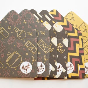 May include: A set of six decorative envelopes with various patterns and designs. The envelopes feature illustrations of candles, potions, hourglasses, and geometric shapes in gold and white on a dark brown background. Each envelope has a "thank you" sticker.