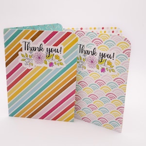 May include: Two folded greeting cards with the text "Thank you!" and floral illustrations. One card has diagonal stripes in various colors, while the other features a wave pattern in pastel shades. The cards are designed for expressing gratitude.