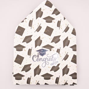May include: A graduation card with a pattern of brown mortarboards on a white background. A sticker with a blue graduation cap and the word "Congrats" is in the center. The card is shaped like a house.