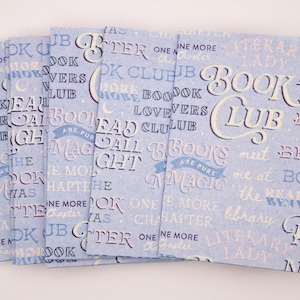 Może przedstawiać: Stos jasnoniebieskich książek w miękkiej oprawie ze słowami "Book Night Club" w stylu vintage. Okładki są wzorzyste ze słowami związanymi z książkami w różnych czcionkach i kolorach, tworząc motyw literacki.