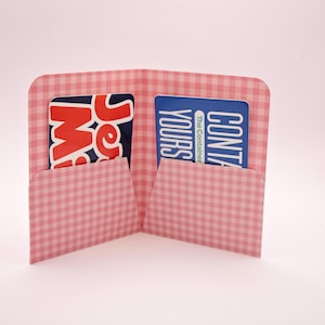 Könnte beinhalten: Eine rosa-weiß karierte Geldbörse mit zwei Fächern. Jedes Fach enthält eine Karte. Eine Karte hat roten und weißen Text mit der Aufschrift "Je M". Die andere Karte hat blauen und weißen Text mit der Aufschrift "Yours The Container".