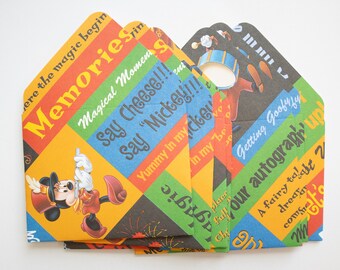 May include: A stack of colorful, patterned paper envelopes featuring Mickey Mouse and Goofy illustrations. The envelopes have diagonal stripes with phrases like "Memories" and "Say Cheese!!!" printed on them.