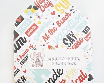 May include: A white envelope with a colorful pattern of words and phrases like "Happy Day" and "Vacation." A label reads "MOUSEKEEPING, THANK YOU" with a cartoon castle and mouse ears. The envelope is shaped like a house.