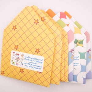 May include: A stack of paper envelopes featuring a yellow grid pattern with red stars and a white rectangular sticker. The sticker displays colorful cartoon characters and the text: To infinity and beyond! Thank you for keeping our room toy-riffic & making our stay extra magical!