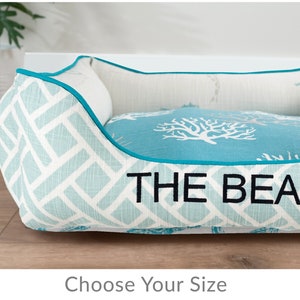 May include: A turquoise and white dog bed with a diamond pattern and a turquoise border. The bed has a white coral and seahorse design and the text "THE BEAN" embroidered on the front.
