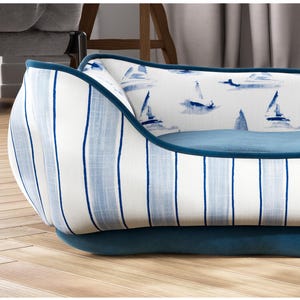 May include: A dog bed with a nautical theme. The bed features a blue and white striped exterior with a blue trim. The interior has a blue cushion and a white fabric with blue sailboat illustrations. The bed sits on a wooden floor.