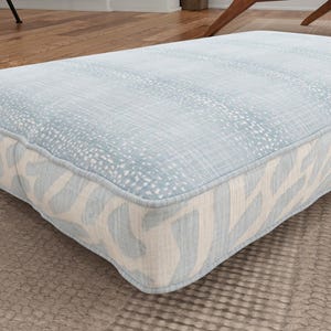 May include: A rectangular floor cushion featuring a light blue top surface with a white speckled pattern and sides decorated with a repeating light blue and cream abstract organic shape design, finished with piped edges along the top perimeter.