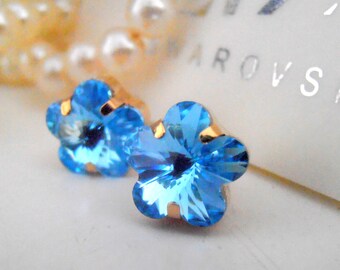 May include: A pair of blue flower-shaped earrings with gold settings. The earrings are on a white surface with a white and gold background.