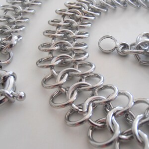 May include: A silver chain necklace with a large, round clasp. The chain is made of interlocking metal rings.
