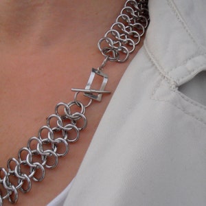 May include: A silver chain necklace with a square clasp. The necklace is made of small, interlocking metal rings.