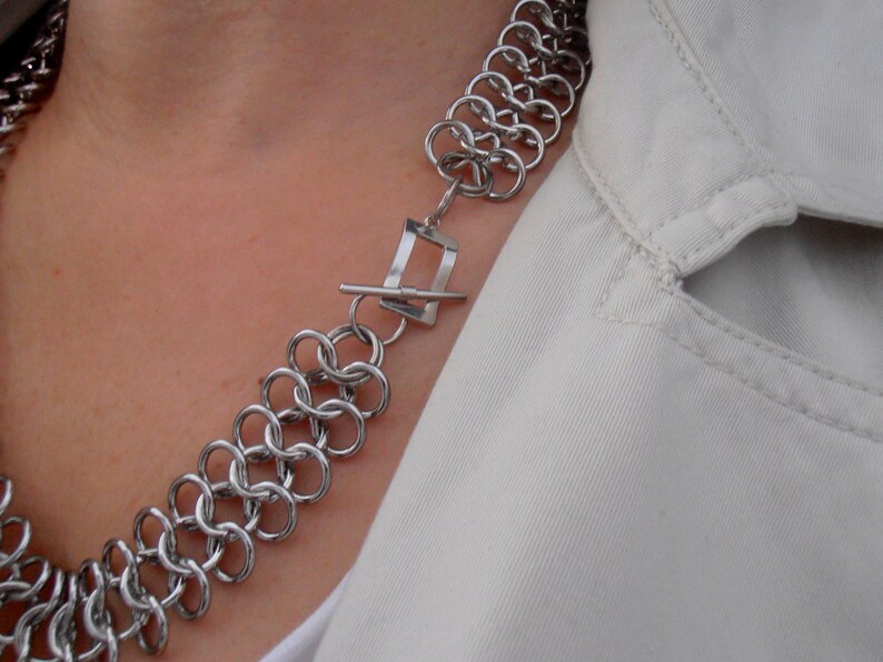 May include: A silver chain necklace with a square clasp. The necklace is made of small, interlocking metal rings.