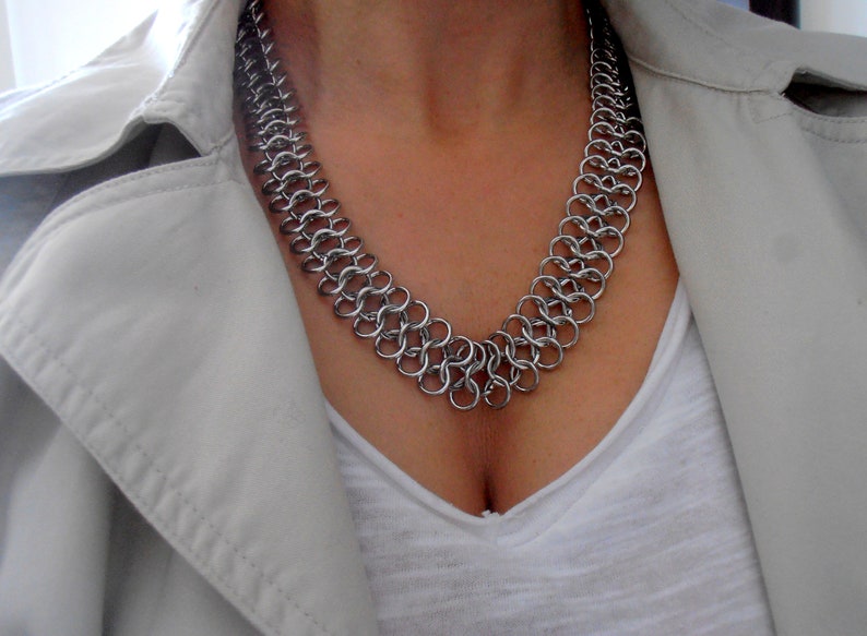 May include: A silver chainmail necklace with a V-shaped design. The necklace is worn by a person with a white shirt and a beige jacket.