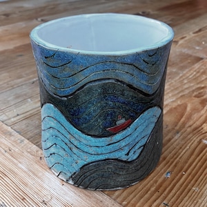 May include: A ceramic cylindrical vase with a nautical theme. The vase features a design of blue and dark teal waves with a small red boat. The interior is white, and the vase sits on a wooden surface.