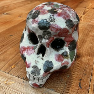 May include: A ceramic skull with a white base, adorned with red, black, and green polka dots. The skull features hollow eye sockets and a detailed jawline. This decorative piece is suitable for home decor.
