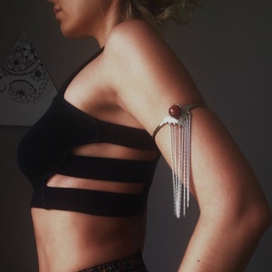 May include: A woman wearing a black crop top with cutouts and a silver arm cuff with a red stone. She is also wearing a patterned skirt and bracelets.