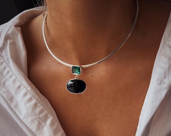 May include: A silver-toned choker necklace featuring a square-cut emerald green stone pendant connected to a larger, smooth, oval-shaped black stone pendant. The necklace rests against a white collared shirt, showcasing a metallic finish.