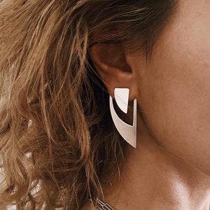 May include: A woman with curly blonde hair wearing a black mesh tank top and silver geometric earrings.