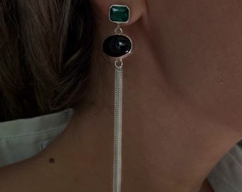 May include: A silver drop earring featuring a rectangular emerald green stone at the top, a smooth oval black stone in the center, and a long, delicate silver chain tassel hanging from the bottom of the oval stone.