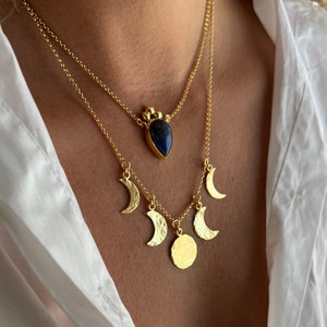 Puede incluir: Un collar de oro con un colgante de piedra azul y cinco dijes de media luna dorada. El collar está en capas y tiene una cadena delicada.