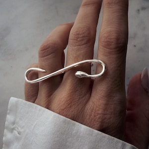 Può includere: Un anello color argento dal design unico e allungato. L'anello si curva su due dita, creando un look elegante e moderno. L'anello è indossato sopra una camicia bianca.