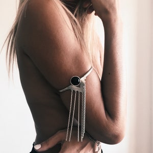 May include: Silver arm cuff with a black stone accent and a chain fringe. The cuff is worn on the upper arm.