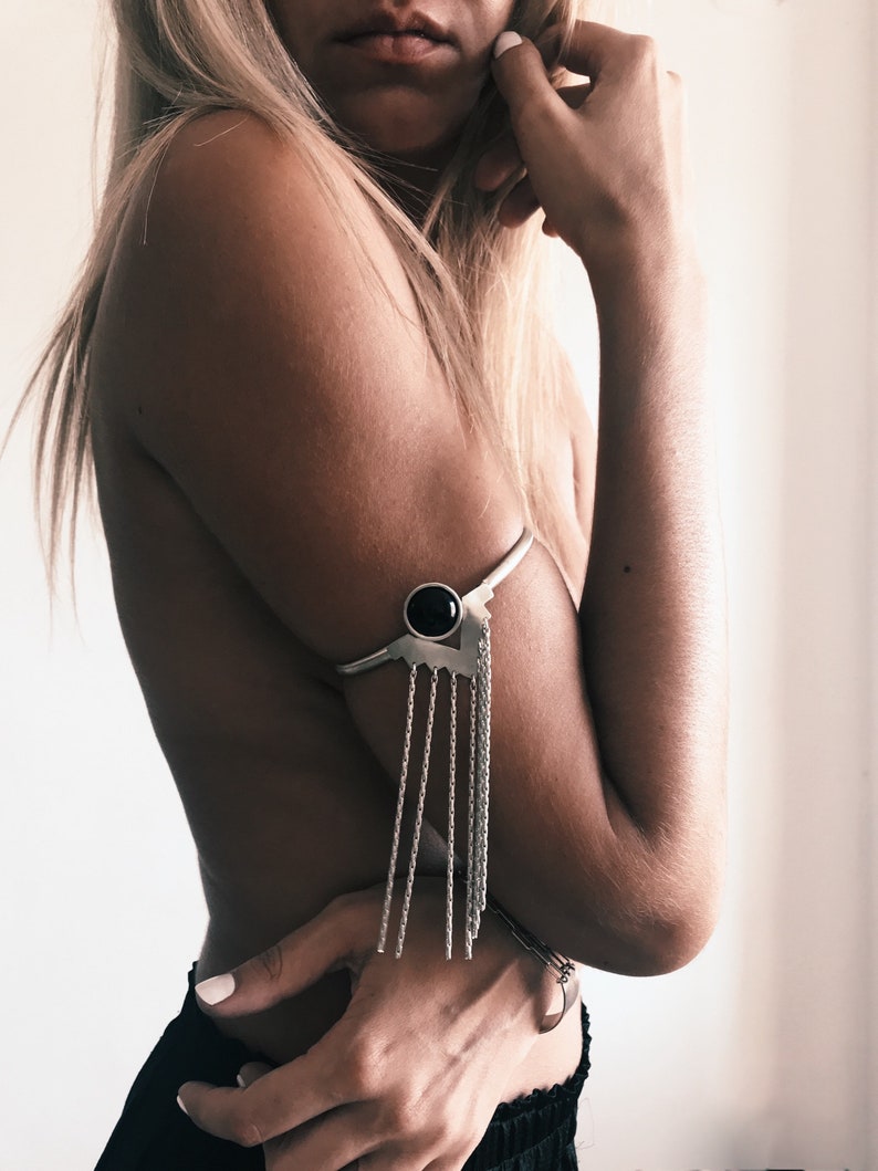 May include: Silver arm cuff with a black stone accent and a chain fringe. The cuff is worn on the upper arm.