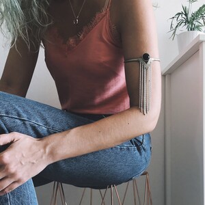 May include: A silver arm cuff with a black gemstone and dangling chains. The cuff is worn on the left arm of a person wearing a pink tank top and blue jeans.