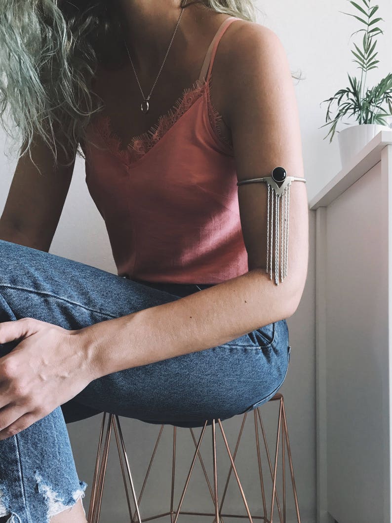 May include: A silver arm cuff with a black gemstone and dangling chains. The cuff is worn on the left arm of a person wearing a pink tank top and blue jeans.