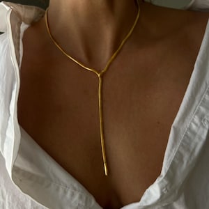 May include: A gold-toned snake chain necklace featuring a Y-shaped lariat design. The necklace consists of a smooth, flexible chain that encircles the neck and extends downwards into a single vertical drop, finished with a sleek, solid metal tip.